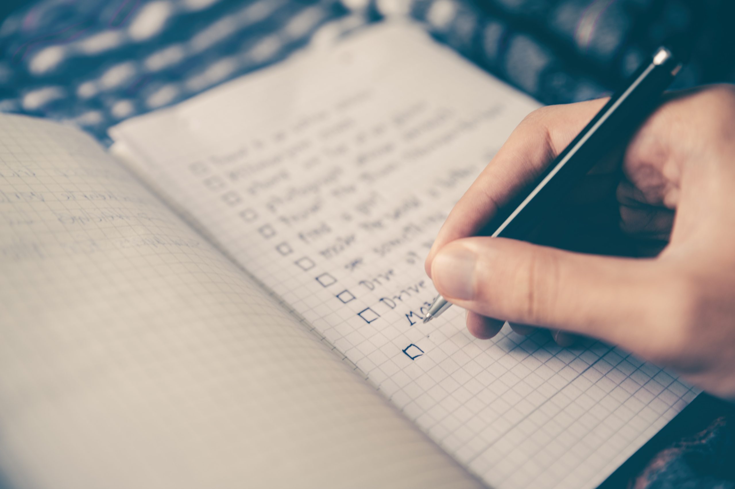A close-up photo of a person writing a checklist in a notebook. The words are out of focus.