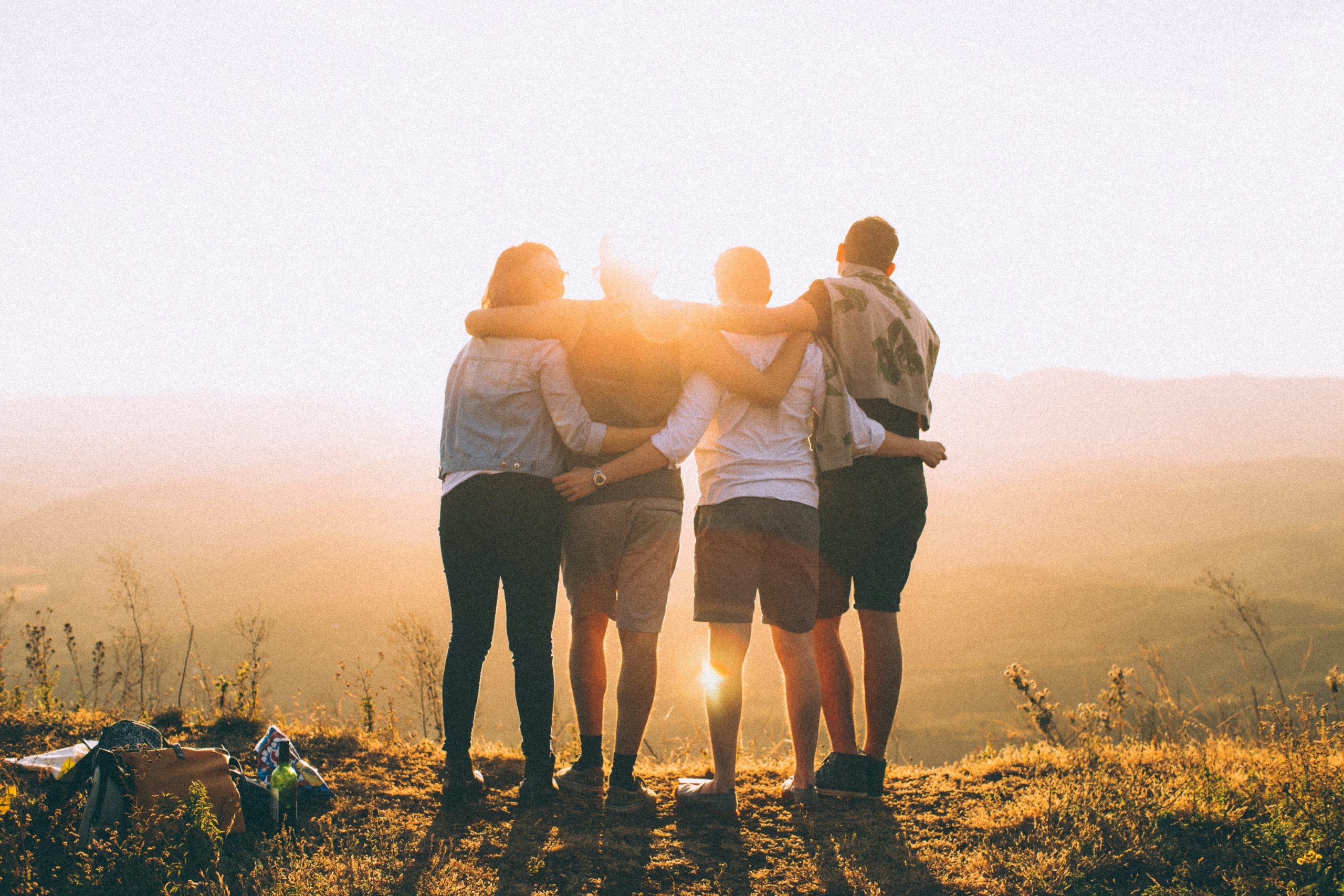 A group of four friends stands with their backs to the camera, backlit by a sunrise.