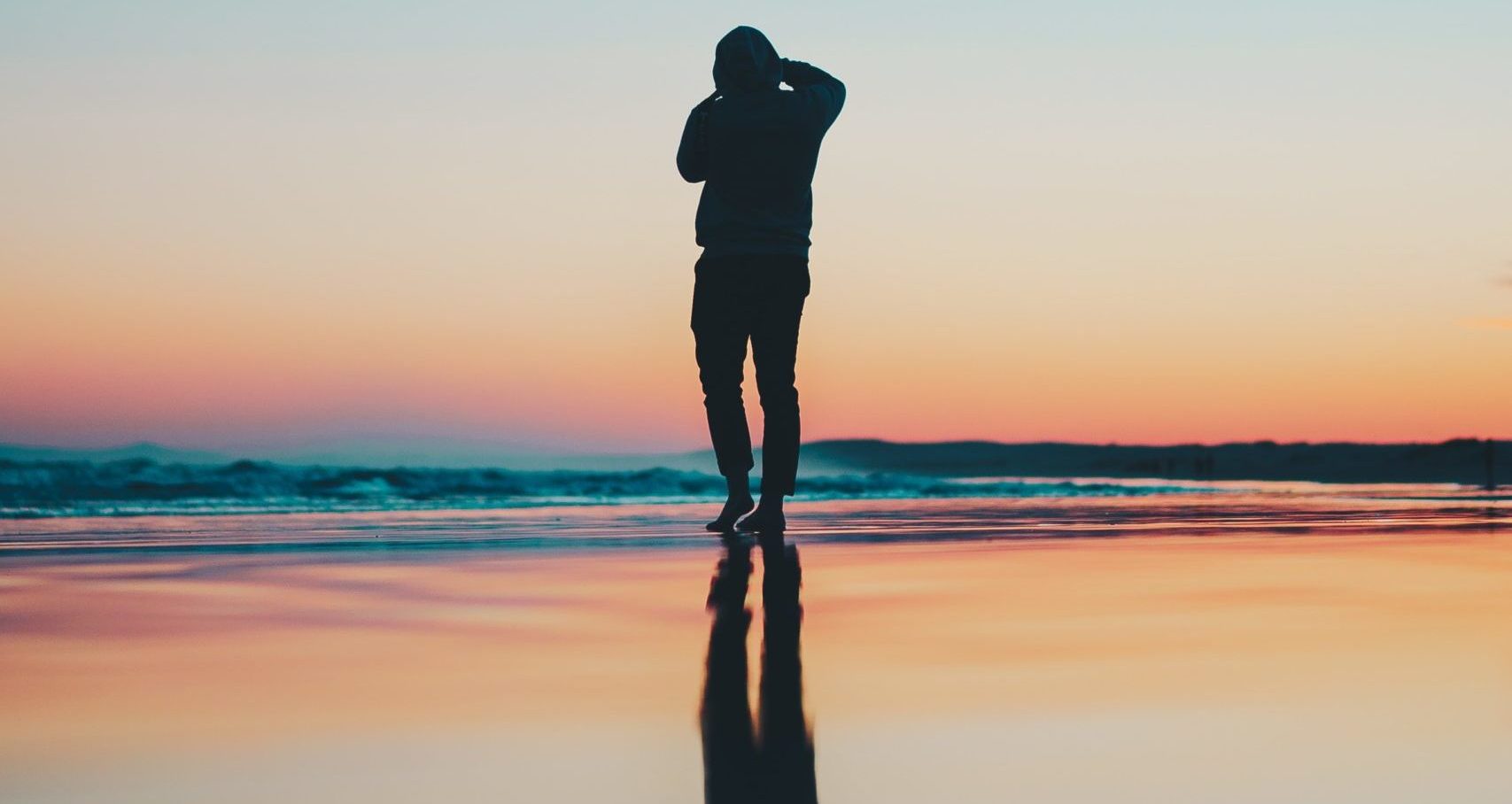 A silhouetted person takes a picture of the ocean as the sun sets. The sunset and the person are reflected in the wet sand.