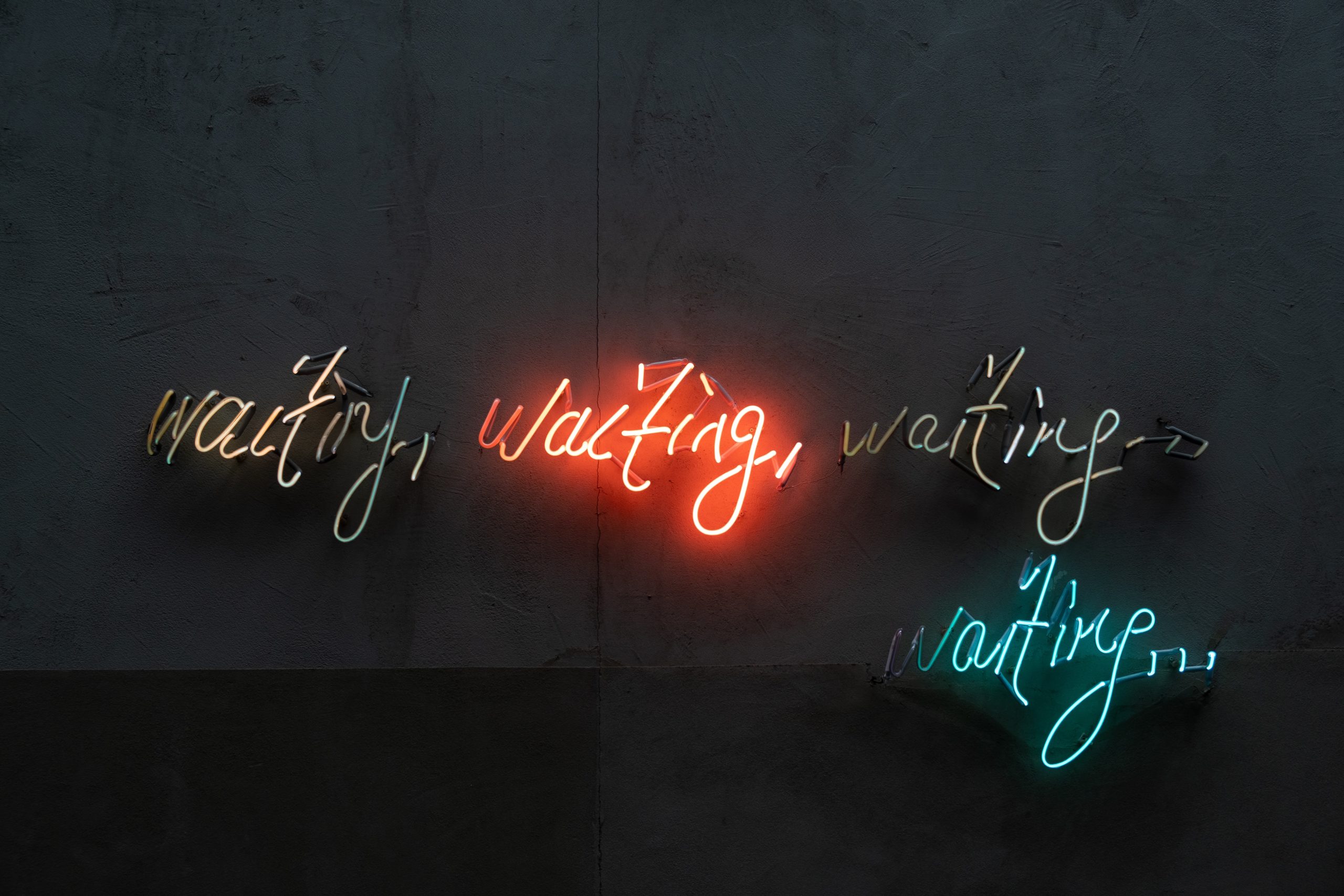 Neon writing on a gray wall spells out "Waiting, waiting, waiting, waiting..." in different colors of red, blue, and white